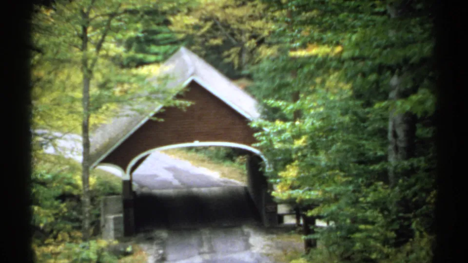 1971 New Hampshire 8mm frame of a brown covered bridge tucked into dense woods, with fall foliage and a road passing through.