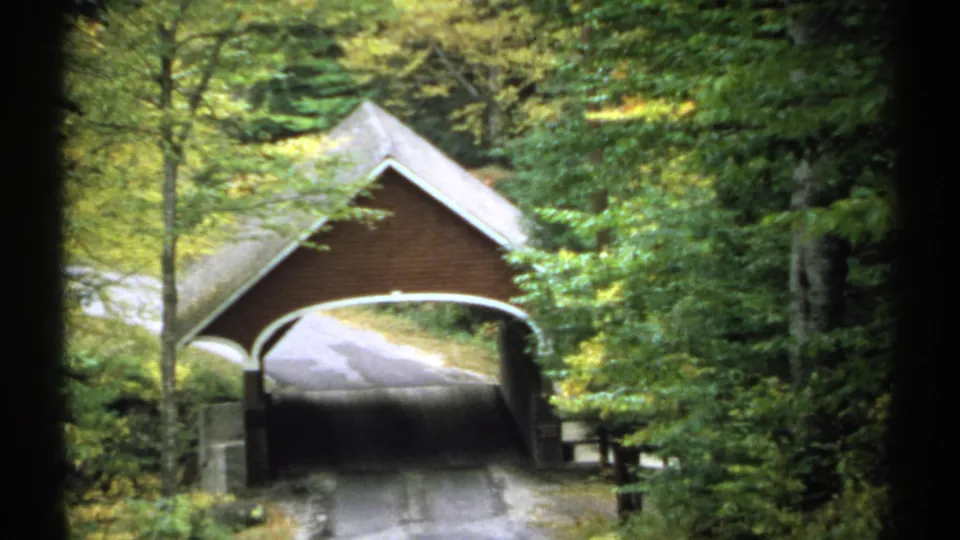 1971 New Hampshire 8mm frame showing a covered bridge on a wooded road, partly framed by dense forest.