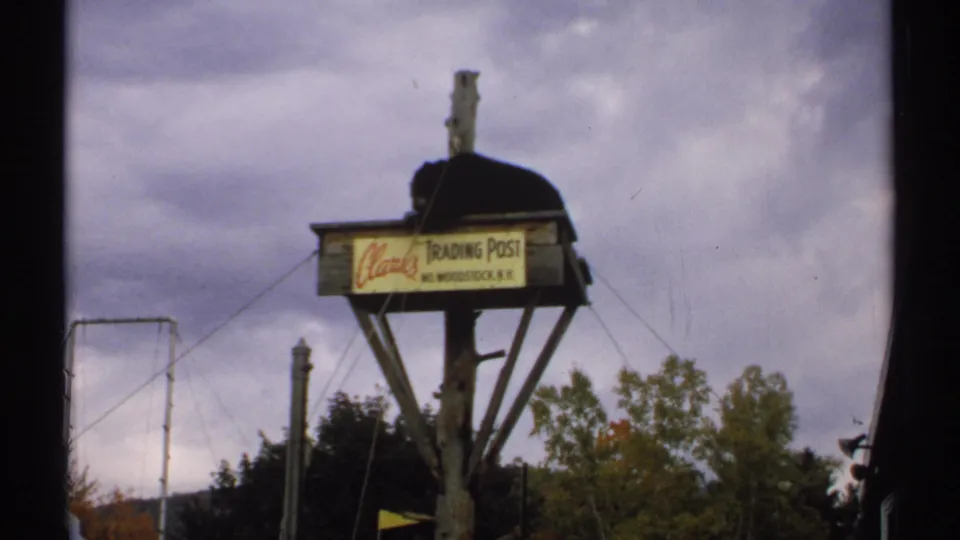 A black bear sits atop the Clarks Trading Post sign in Woodstock, New Hampshire, with trees and poles behind it.