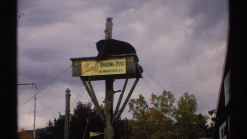 1971 New Hampshire home-movie frame showing a bear on a raised wooden platform with Clark's Trading Post signage and trees nearby.