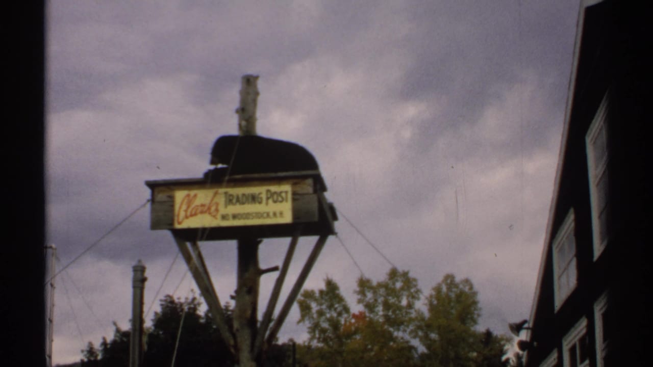 1971 New Hampshire 8mm frame showing the Clarks Trading Post sign, a dark building edge, trees, wall, and chain-link fence.