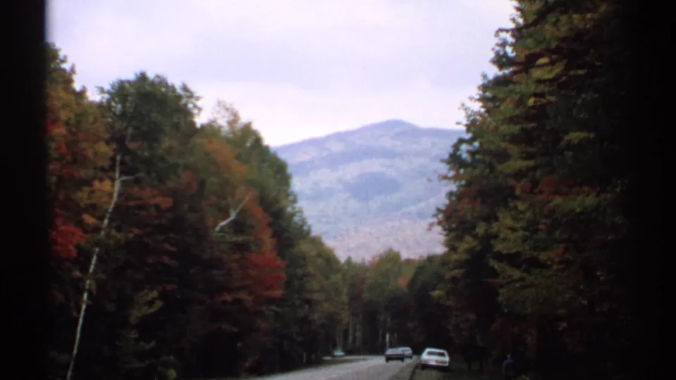 1971 New Hampshire road scene with cars traveling through fall foliage toward a distant mountain under overcast sky.