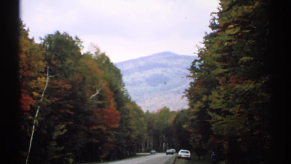 New Hampshire 8mm travel frame showing classic cars on a forest road with fall color and a distant mountain view.