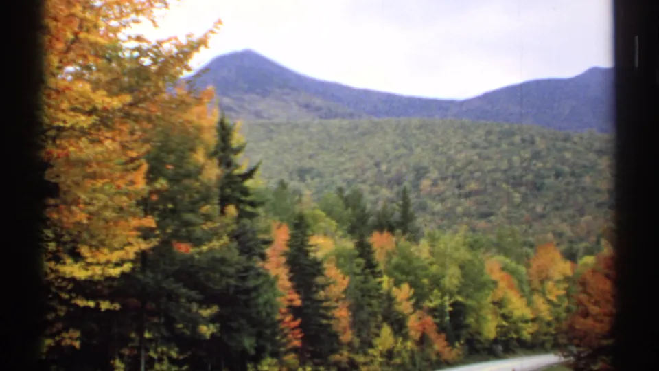 1971 New Hampshire home-movie frame showing orange foliage, dark evergreens, forested hills, and a distant mountain peak.