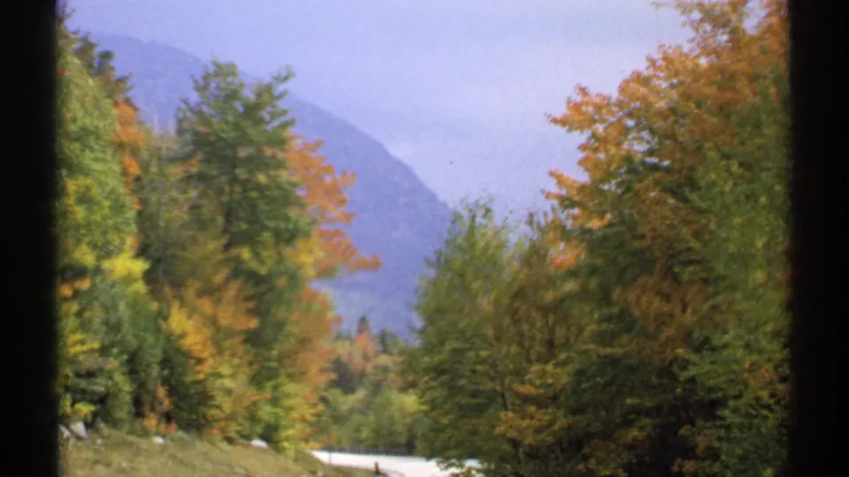 1971 New Hampshire 8mm frame showing a pale road curving past autumn foliage with hazy mountains in the background.