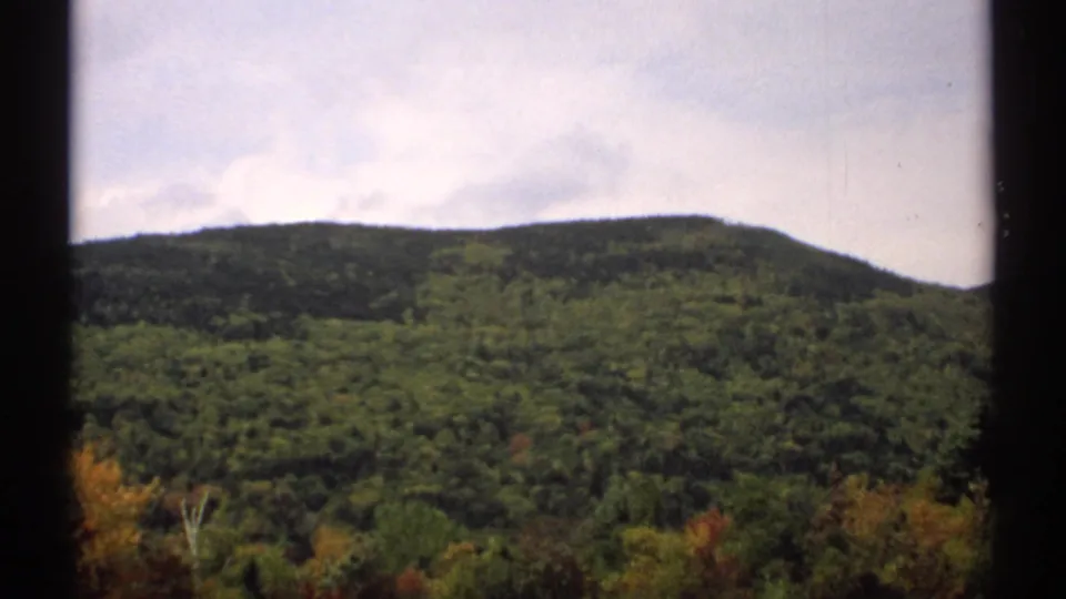 1971 New Hampshire landscape frame showing a wooded mountain slope, cloudy sky, and early autumn color in the lower trees.
