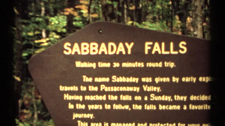 1971 New Hampshire home-movie frame of a Sabbaday Falls sign noting walking time and name-origin text, set in forest shade.