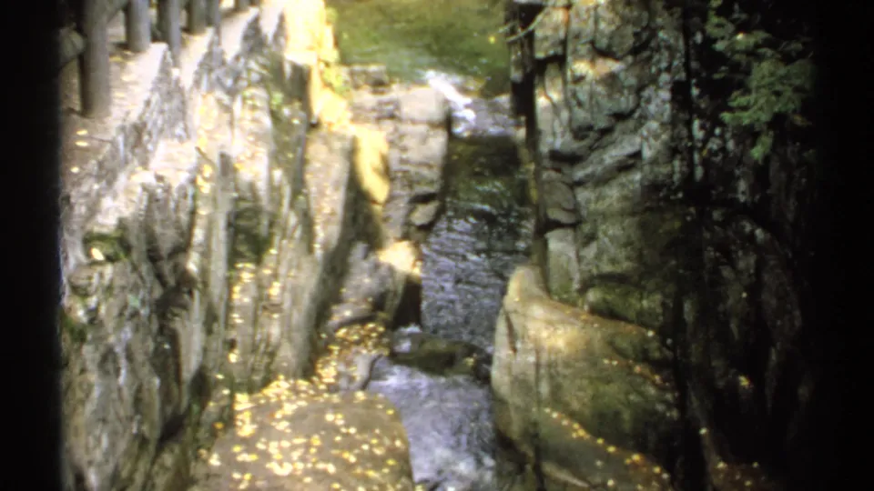 1971 New Hampshire 8mm frame showing the Sabbaday Falls rocky gorge from a visitor overlook, with water below and dark stone walls around it.