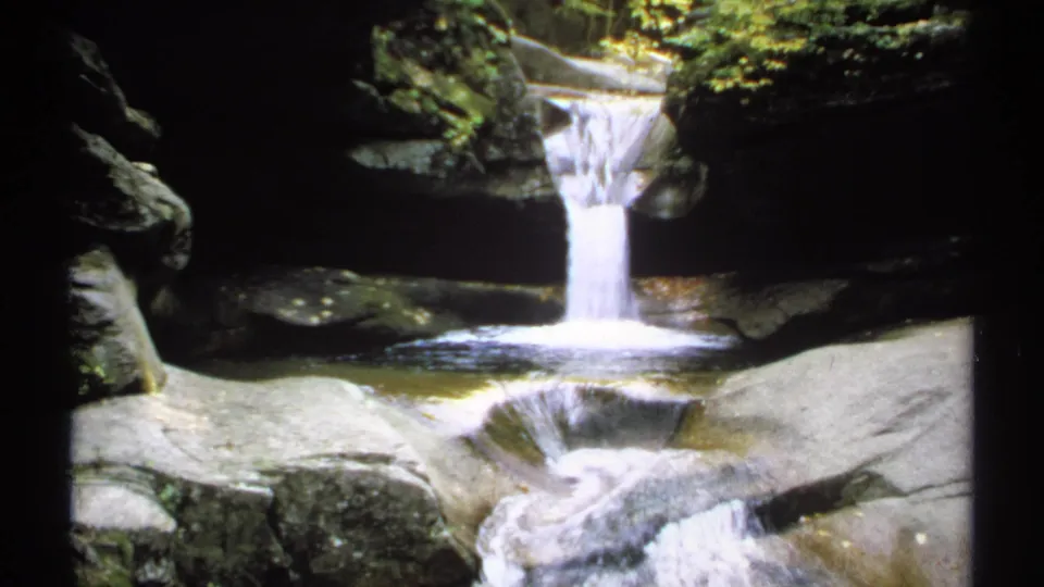 1971 New Hampshire 8mm frame showing the same rocky stream with a small cascade, pale stone shelves, and deep gorge shadows.