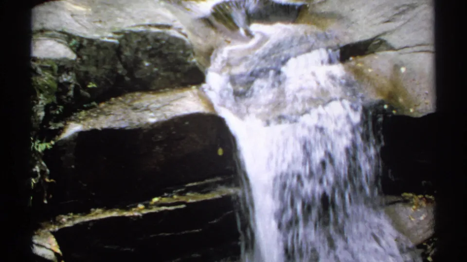 1971 New Hampshire home-movie frame with white water rushing over smooth layered rocks in a shaded gorge.