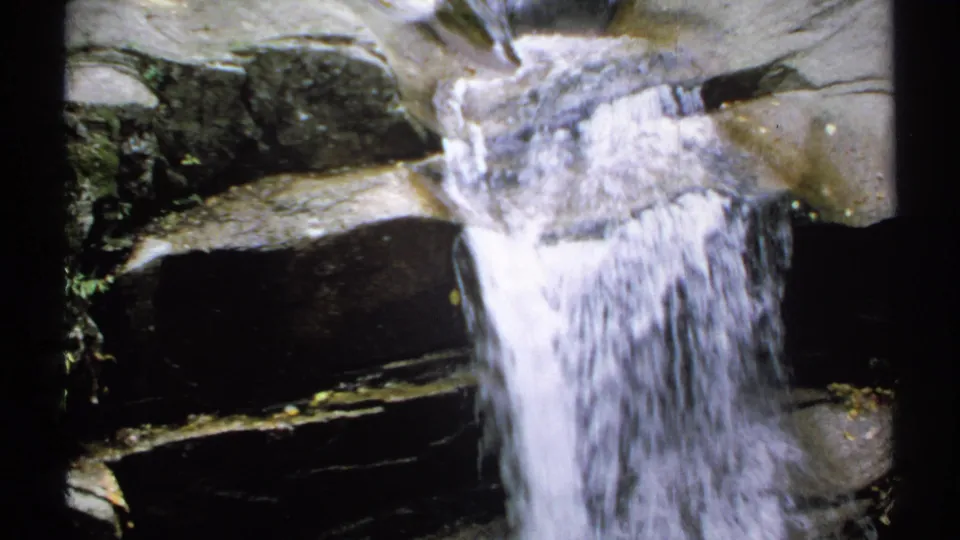 1971 New Hampshire 8mm frame with a narrow sheet of water pouring down uneven rock, framed by dark stone edges.