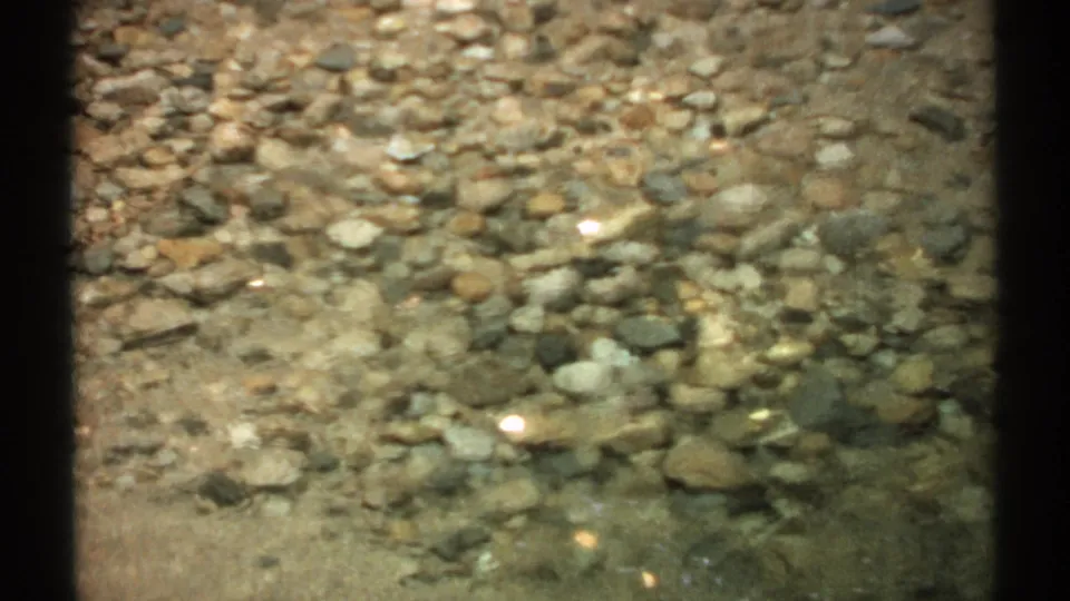 1971 New Hampshire home-movie frame of a shallow stream bed, with stones distorted by moving clear water.