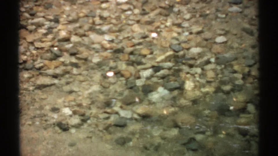 1971 New Hampshire 8mm frame showing pebbles under a clear creek surface with bright sun reflections.