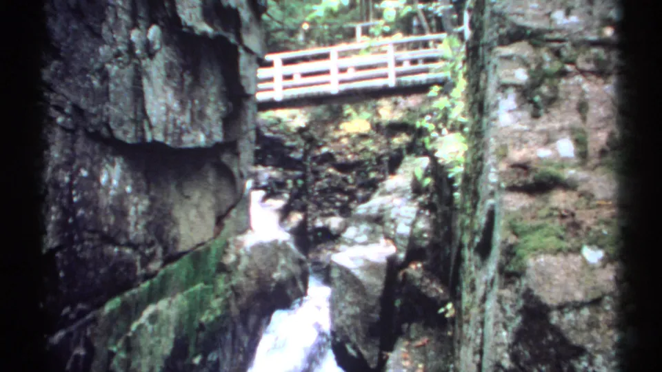 1971 New Hampshire home-movie frame showing a wooden bridge above a mossy rock gorge and rushing stream water.