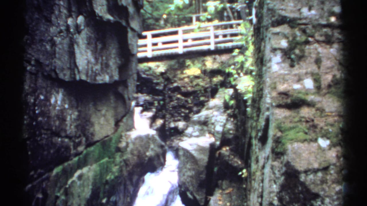 1971 New Hampshire 8mm frame showing a narrow rocky gorge, white water, mossy stone, and a wooden footbridge above.
