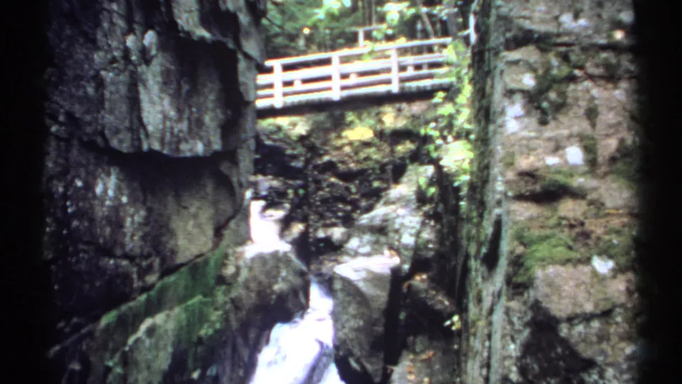 1971 New Hampshire 8mm frame with a wooden footbridge crossing above a shadowed gorge and falling water.