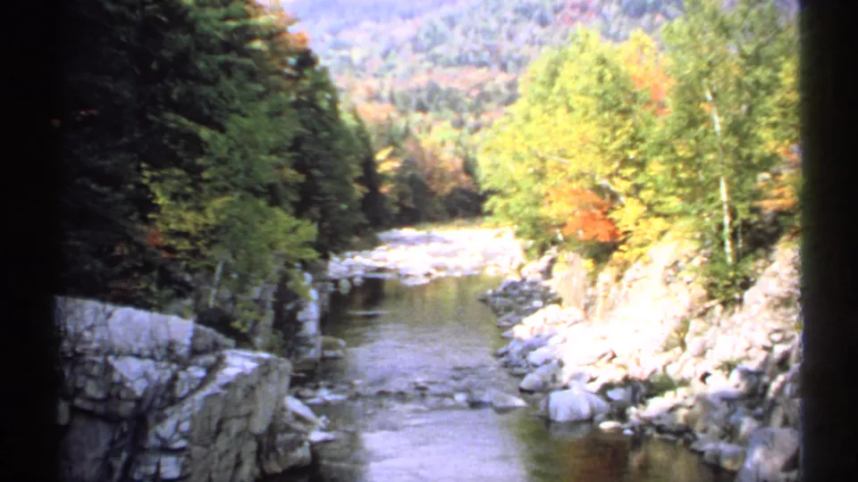 1971 New Hampshire 8mm frame showing a shallow river through a forested valley with boulders and early autumn color.