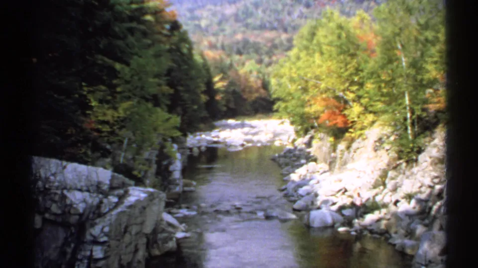 1971 New Hampshire 8mm frame showing a calm creek, pale rocks, dense trees, and early autumn color in a wooded gorge.