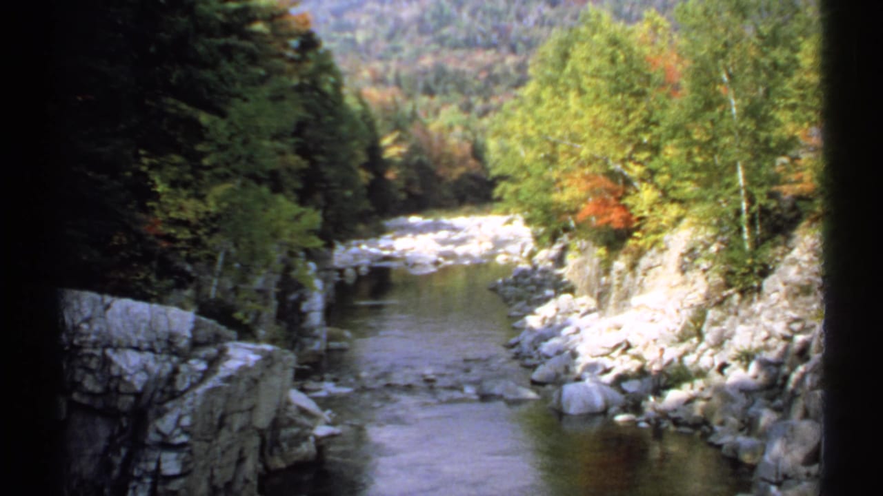 1971 New Hampshire scenic frame with a creek, stone banks, green and orange foliage, and a wooded slope beyond.