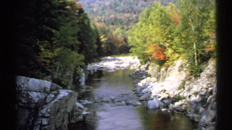 1971 New Hampshire scenic frame with a creek, stone banks, green and orange foliage, and a wooded slope beyond.