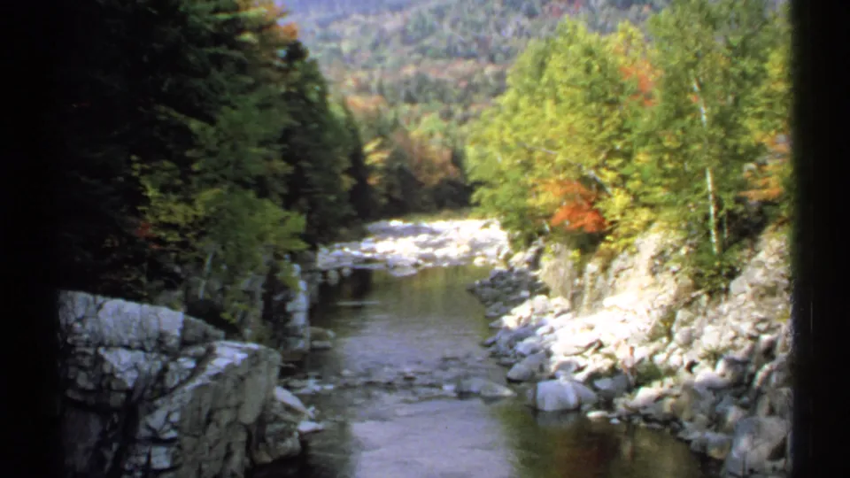1971 New Hampshire 8mm view of a quiet creek, rocky edges, dense woodland, and muted autumn foliage.