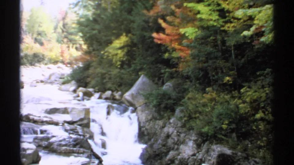 1971 New Hampshire frame showing a small waterfall rushing over boulders beside thick forest and autumn foliage.