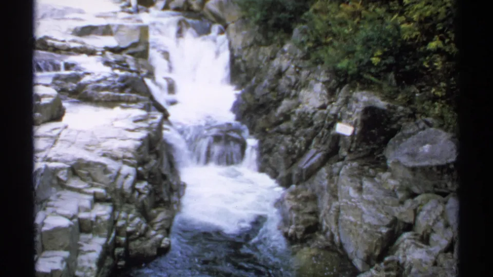 1971 New Hampshire 8mm frame of a forest waterfall and rocky stream, with boulders, dense foliage, and a small sign-like marker visible near the rocks.