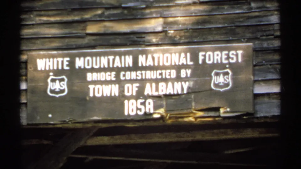 1971 8mm close-up of the White Mountain National Forest bridge marker with U.S. Forest Service shields and Town of Albany text.