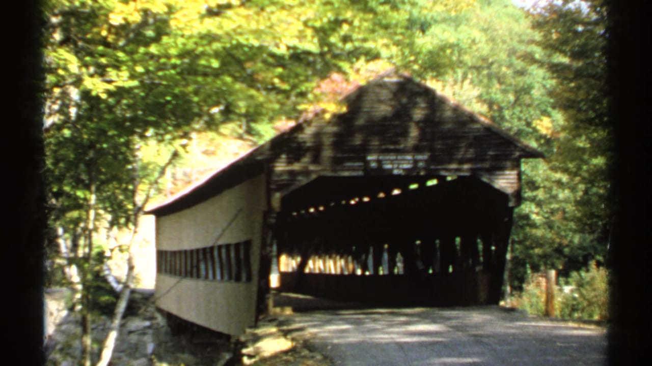 Wooden covered bridge ahead on a wooded road with green and yellow foliage.