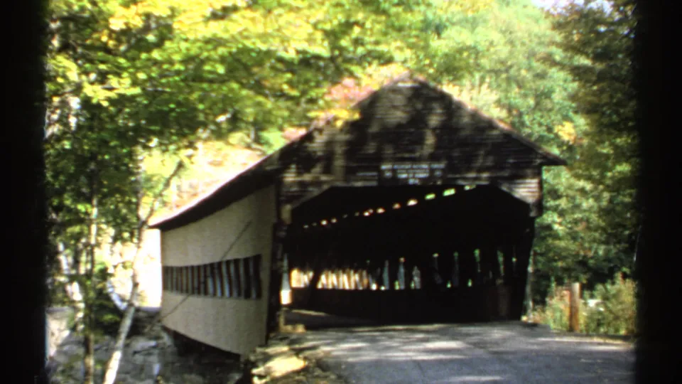 A closer 1971 home-movie view shows the covered bridge opening, roofline, side wall, and wooded New Hampshire roadside.