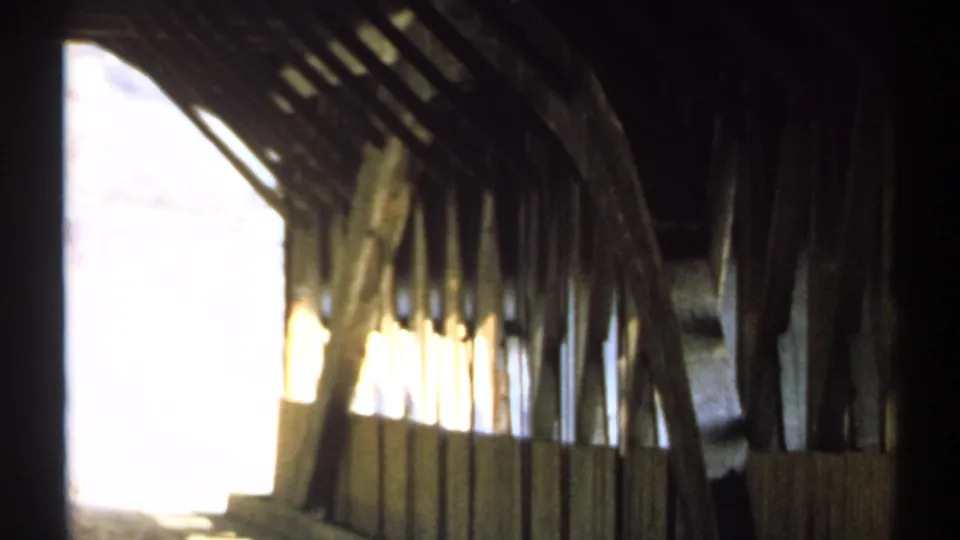 Inside the covered bridge, the 8mm frame shows dark wooden trusses, side openings, and bright exterior light spilling into the structure.