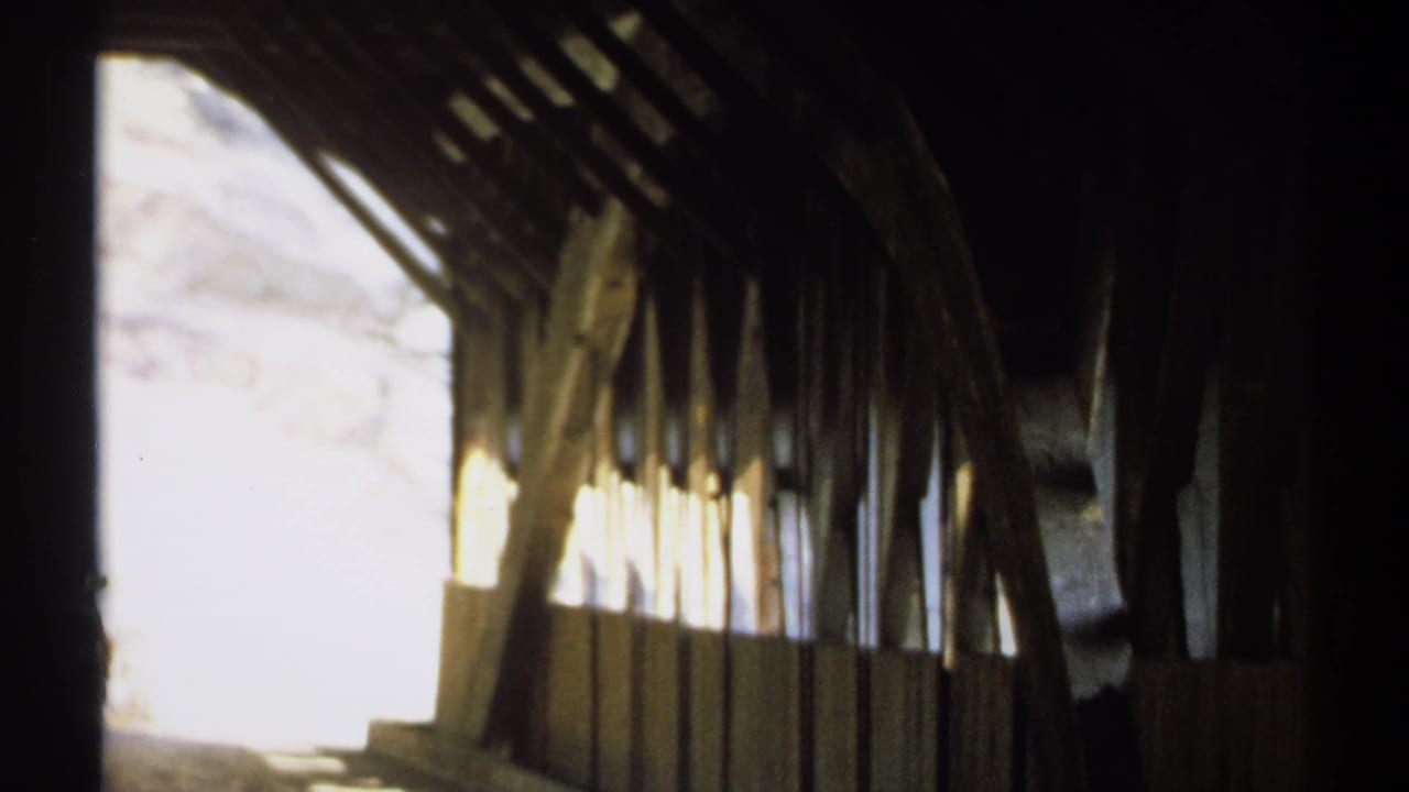 Covered bridge interior with dark rafters and a bright exterior opening.