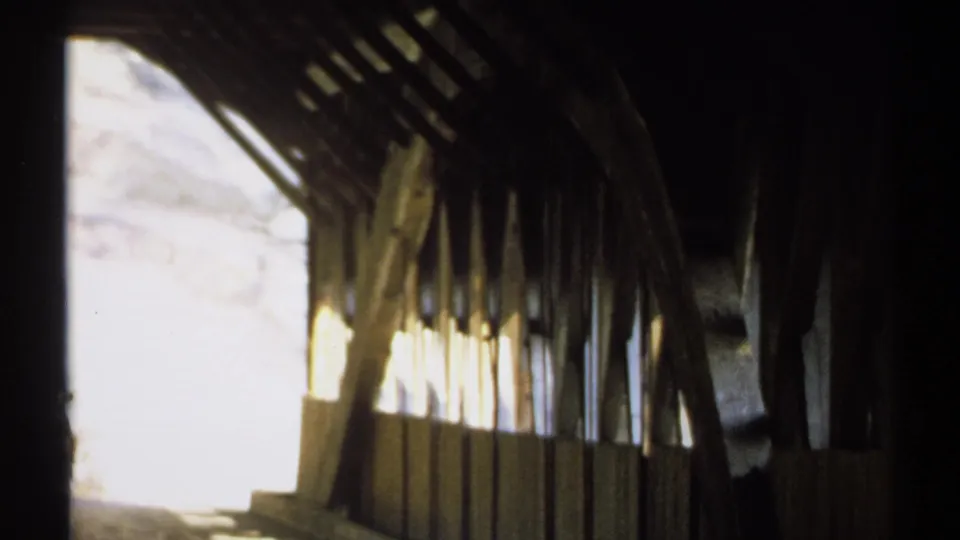 Inside the New Hampshire covered bridge, the camera faces along wooden trusses toward a blown-out daylight opening.