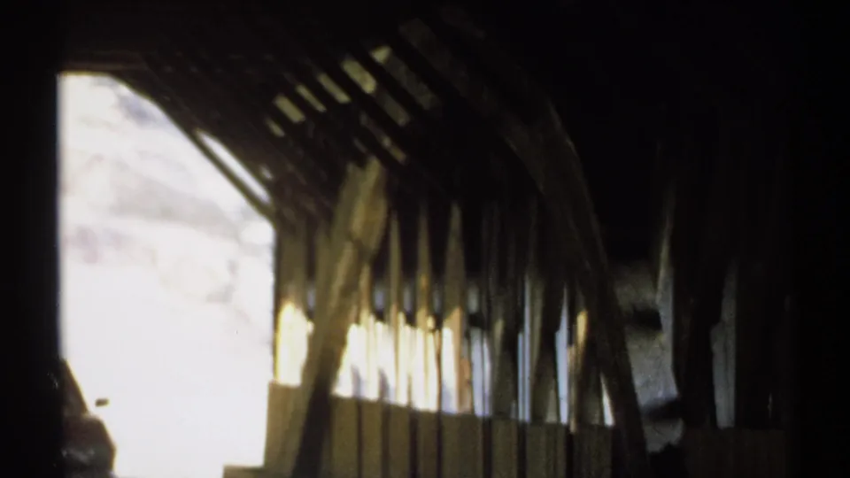 Handheld 8mm frame from inside the covered bridge, with heavy shadow, side openings, and rustic wood structure.