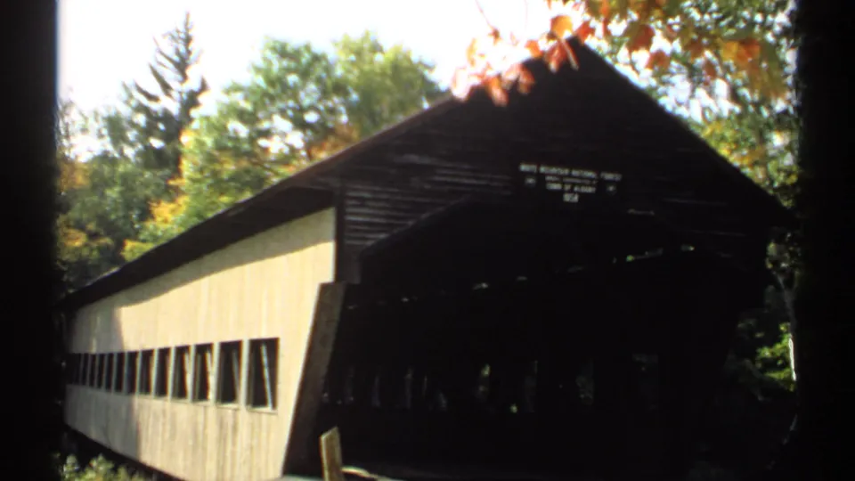 Exterior view of a New Hampshire covered bridge in 1971, with light siding, dark entrance, forest, and autumn leaves.