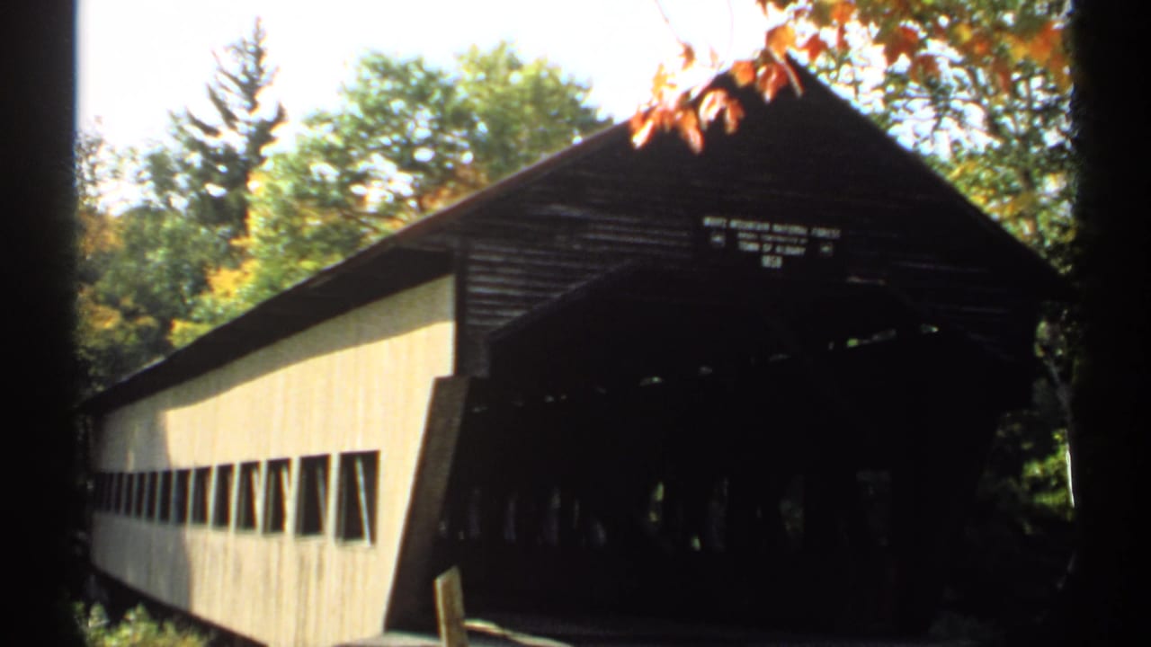 1971 New Hampshire home-movie frame of a covered bridge with pale siding, dark entrance, autumn foliage, and partial public signage.