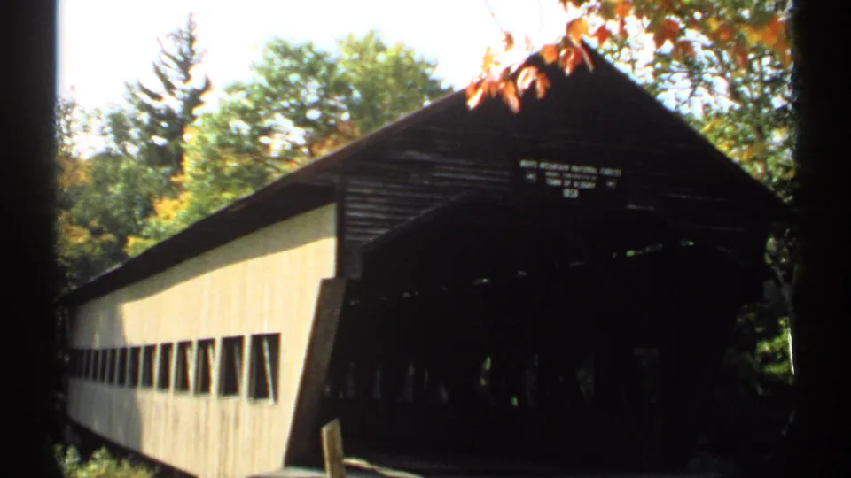 1971 New Hampshire home-movie frame of a covered bridge with pale siding, dark entrance, autumn foliage, and partial public signage.