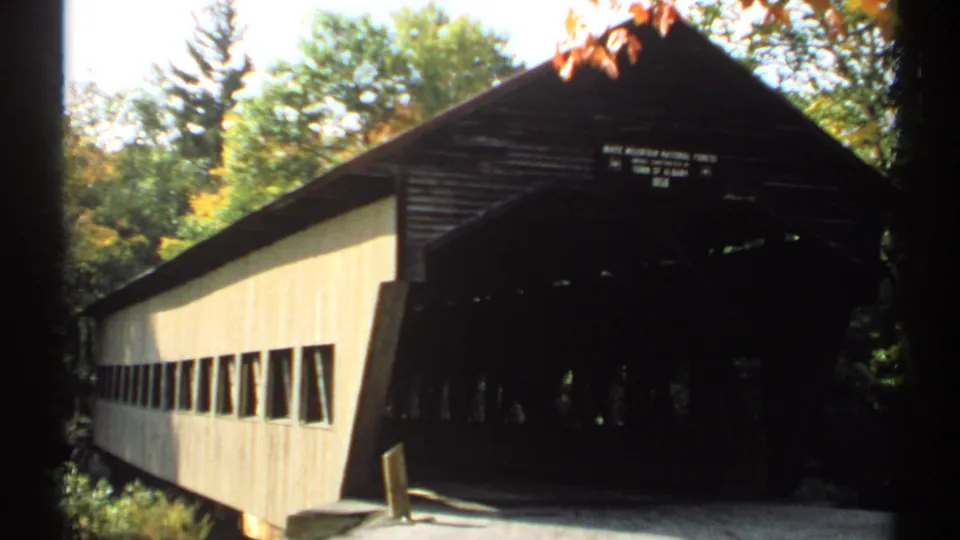 8mm travel frame showing the covered bridge's pale side wall, dark portal, overhead sign, and surrounding autumn foliage.