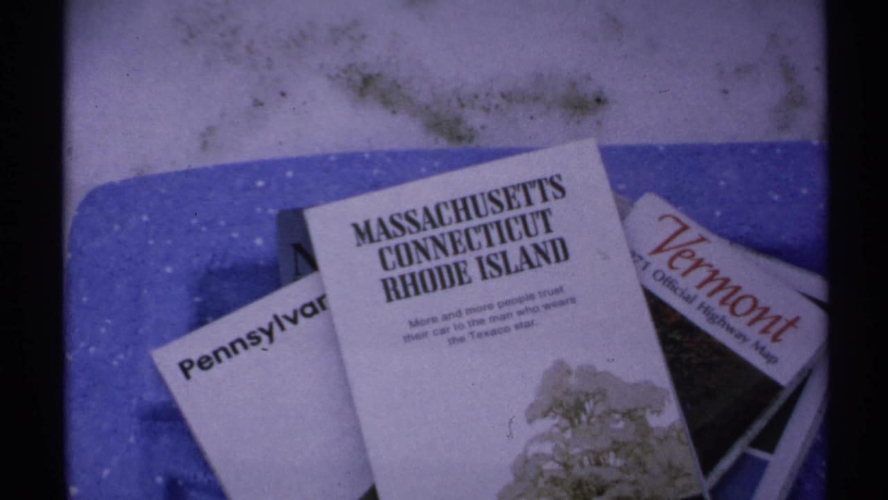 1971 home-movie frame showing state travel brochures on a blue surface, with readable Northeast state names and visible film grain.