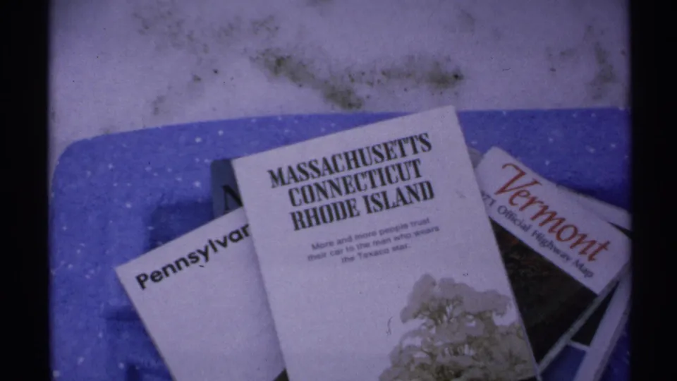 1971 home-movie frame showing state travel brochures on a blue surface, with readable Northeast state names and visible film grain.