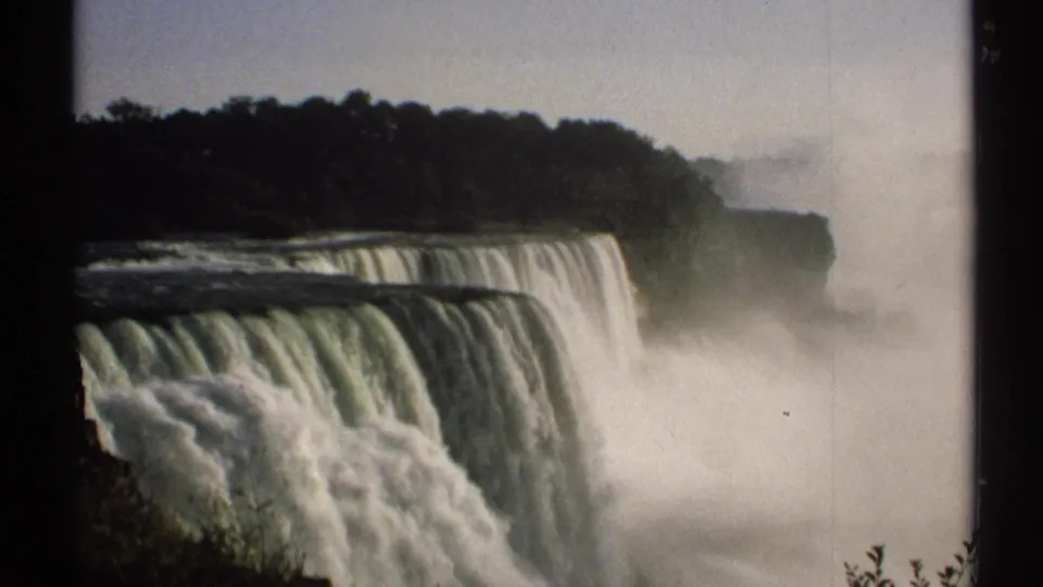 8mm New York travel frame showing a side view of a broad waterfall with mist, dark trees, and a cliff edge in the distance.