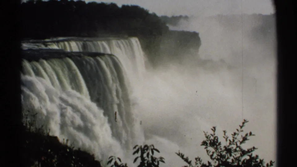 8mm footage of Niagara Falls from a visitor viewpoint, showing cascading water, spray, and silhouetted foliage in front.