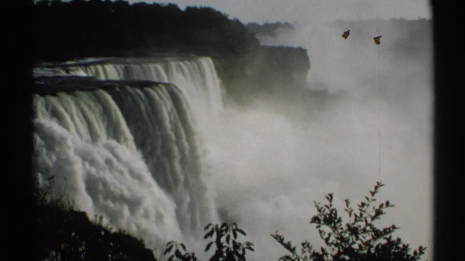 1971 home-movie frame of Niagara Falls in New York, with white spray filling the lower view and shrubs at the overlook edge.