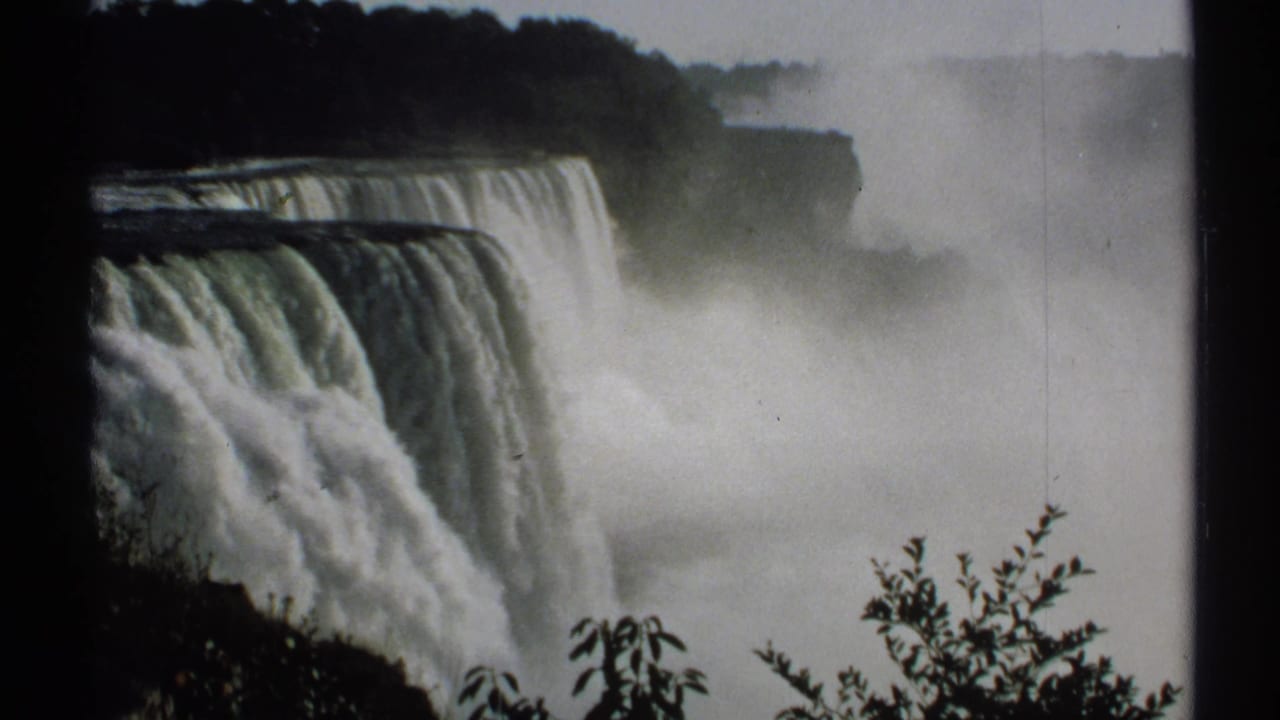 1971 8mm close view of Niagara Falls from New York, with water dropping over the crest and mist rising around dark shoreline foliage.