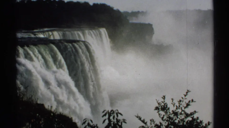 1971 8mm close view of Niagara Falls from New York, with water dropping over the crest and mist rising around dark shoreline foliage.