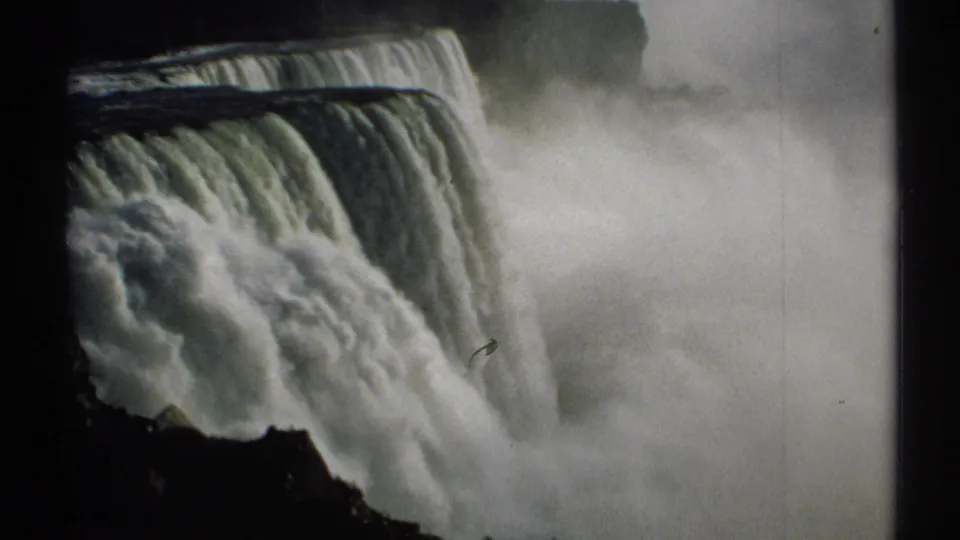 Close 1971 home-movie frame of Niagara Falls, showing the cascade, heavy spray, distant tree line, and dark rock edge.