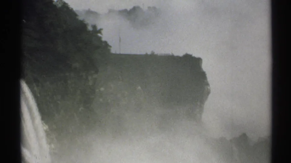 1971 New York 8mm frame showing a misty waterfall falling beside a dark tree-covered cliff, with heavy spray softening the view.
