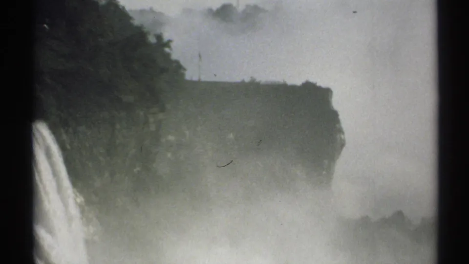 1971 New York home-movie frame with a waterfall at the left edge, a wooded cliff across the center, and mist filling the lower scene.