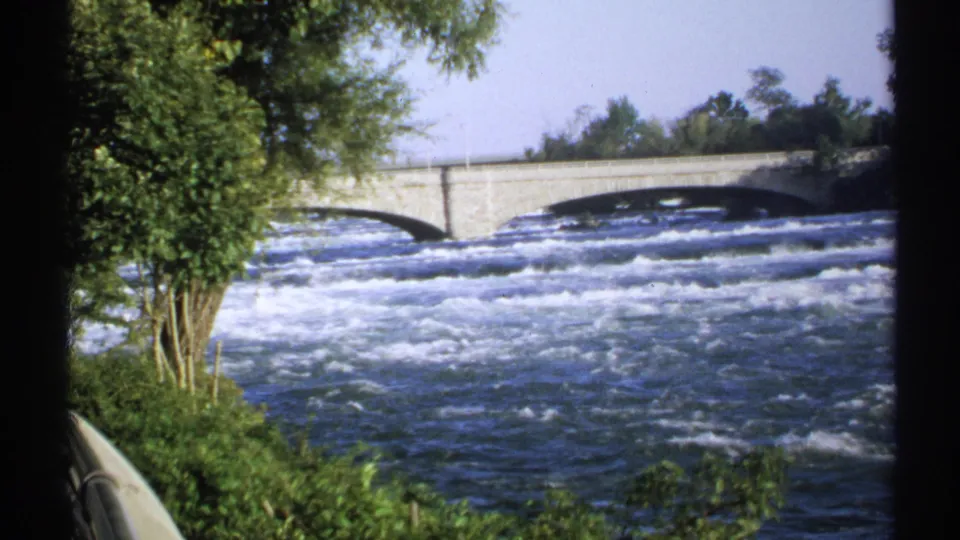 1971 New York 8mm frame showing white-water rapids moving beneath a stone arch bridge, with trees and greenery along the riverbank.