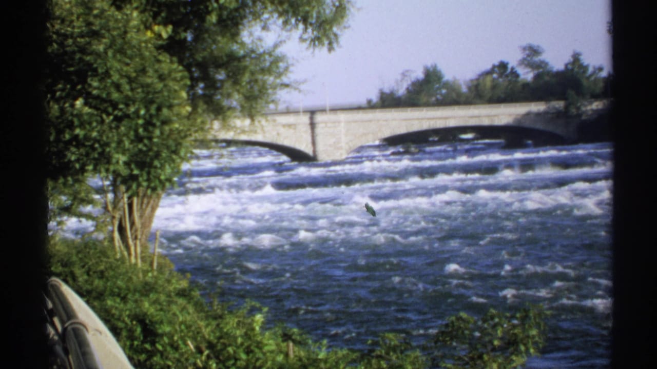 Fast whitewater river flowing under a stone arch bridge with trees and railings nearby.
